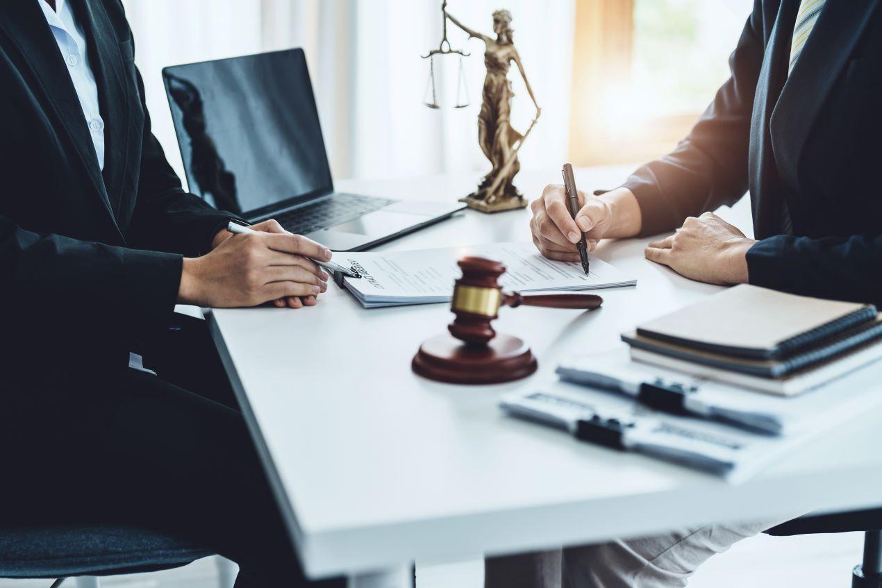 Zwei Personen in einem Büro besprechen Unterlagen. Eine Justitia-Statue und ein Richterhammer symbolisieren rechtliche Unterstützung durch die Kanzlei Ra-Hartung.